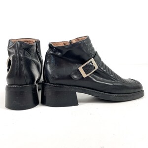May include: Black leather ankle boots with a square toe and a small block heel. The boots feature a silver-tone buckle and a zipper closure. The leather has a slightly worn appearance.