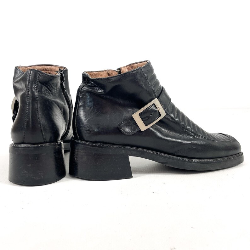 May include: Black leather ankle boots with a square toe and a small block heel. The boots feature a silver-tone buckle and a zipper closure. The leather has a slightly worn appearance.