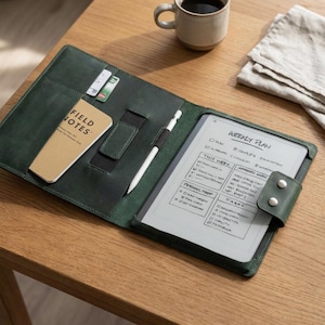 Puede incluir: Un folio de cuero verde oscuro con una tableta, un bolígrafo y un bloc de notas. La tableta muestra un plan semanal. El folio tiene ranuras para tarjetas y un cierre magnético. Una taza de café y una servilleta están sobre la mesa de madera.