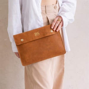 May include: A brown leather laptop sleeve with the initials "BS" in gold lettering. The sleeve has a flap closure secured by two gold-colored snap buttons. The overall design is minimalist and elegant, suitable for carrying a laptop or tablet.