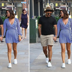 May include: A couple walking down the street, holding hands. The man is wearing a black t-shirt, khaki shorts, a brown hat, and white sneakers. The woman is wearing a blue and white striped dress, a brown hat, and white sneakers.