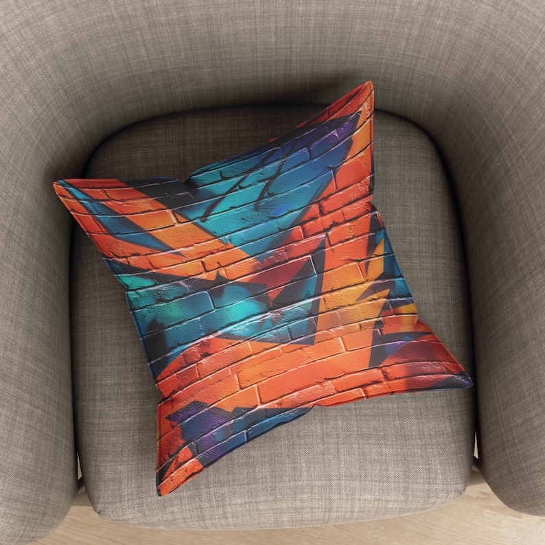 Op de afbeelding: Vierkant sierkussen met een levendig graffiti-achtig ontwerp. Het kussen heeft een bakstenen muurpatroon in tinten oranje, blauw en paars. Het kussen ligt op een grijze fauteuil.
