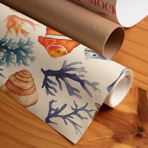 Op de afbeelding: Gerolde kunstdruk met een aquarelontwerp van zeeleven, waaronder een oranje clownvis, blauw koraal en een zeeschelp. De print staat op een beige achtergrond. Een bruine kartonnen koker is zichtbaar op de achtergrond met het woord "MOCK" erop gedrukt.
