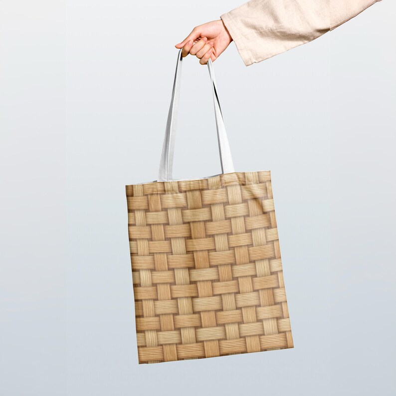 May include: A rectangular tote bag with a woven basket pattern in shades of tan and brown. The bag has white straps and is held up by a person's hand.