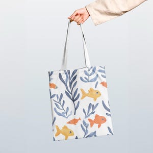 Op de afbeelding: Witte draagtas met een herhalend patroon van gele en oranje vissen, blauw zeewier en bladeren. De tas heeft witte handvatten. Het ontwerp is gedrukt op een witte achtergrond. De tas wordt vastgehouden.