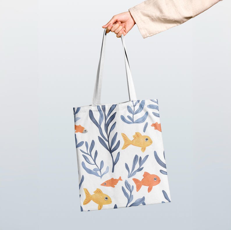 Op de afbeelding: Witte draagtas met een herhalend patroon van gele en oranje vissen, blauw zeewier en bladeren. De tas heeft witte handvatten. Het ontwerp is gedrukt op een witte achtergrond. De tas wordt vastgehouden.