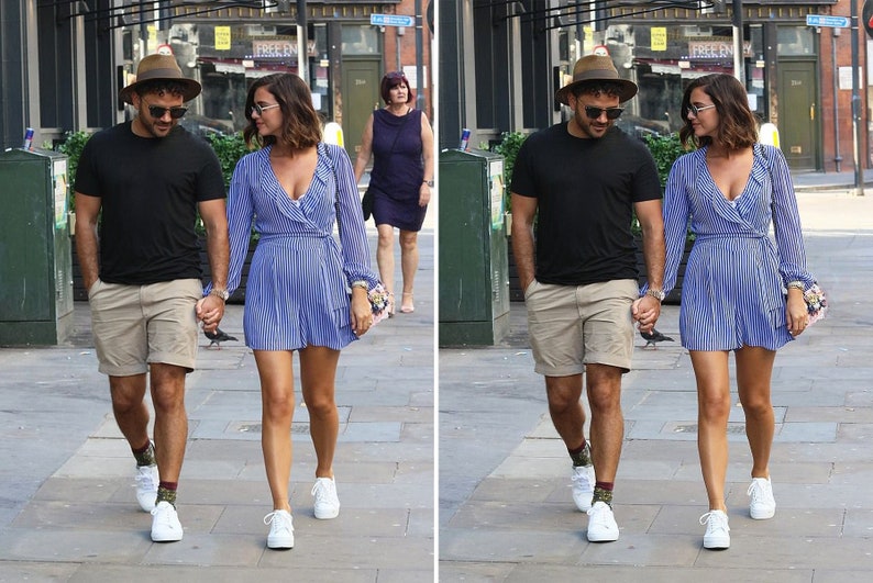 May include: A couple walking down the street, holding hands. The man is wearing a black t-shirt, khaki shorts, a brown hat, and white sneakers. The woman is wearing a blue and white striped dress, a brown hat, and white sneakers.