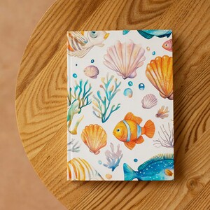 Op de afbeelding: Een hardcover notitieboek met een marien ontwerp. De omslag toont illustraties van oranje en gele vissen, schelpen, koraal en bellen op een witte achtergrond. Het notitieboek ligt op een houten oppervlak.