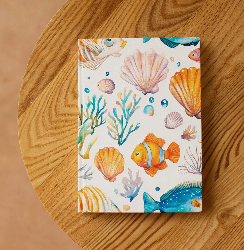 Op de afbeelding: Een hardcover notitieboek met een marien ontwerp. De omslag toont illustraties van oranje en gele vissen, schelpen, koraal en bellen op een witte achtergrond. Het notitieboek ligt op een houten oppervlak.