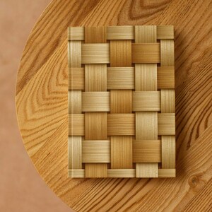 May include: A rectangular notebook with a woven pattern cover in shades of tan and light brown. The notebook is centered on a round wooden table with a light brown finish. The woven pattern gives the notebook a textured appearance.