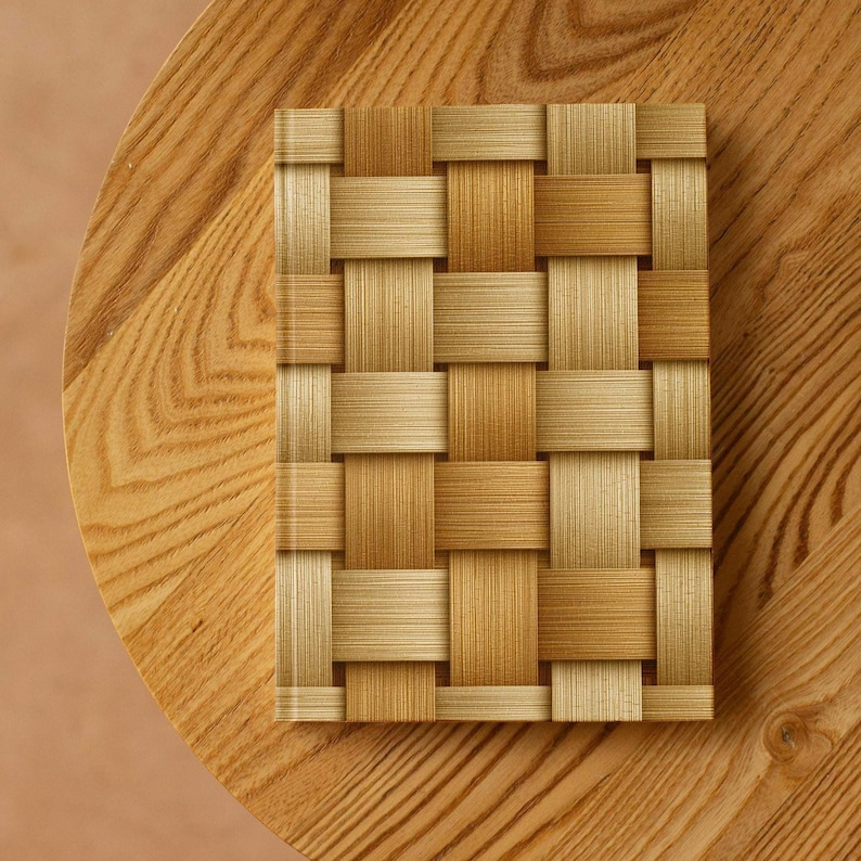 May include: A rectangular notebook with a woven pattern cover in shades of tan and light brown. The notebook is centered on a round wooden table with a light brown finish. The woven pattern gives the notebook a textured appearance.