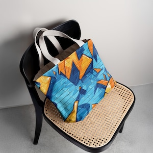 Op de afbeelding: Een tote bag met een blauw baksteenpatroon en oranje en gele geometrische vormen. De tas heeft witte handvatten en een bruine binnenkant. De tas ligt op een zwarte stoel met een geweven zitting.