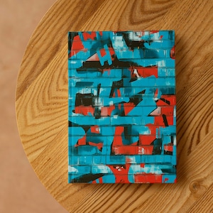 Op de afbeelding: Een rechthoekig notitieboek met een levendig graffiti-achtig ontwerp. De omslag heeft een baksteenpatroon in tinten blauw, rood en zwart. Het notitieboek ligt op een houten oppervlak.