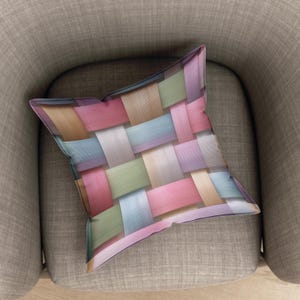 May include: A decorative square pillow with a woven pattern in pastel colors, including pink, blue, green, and beige. The pillow is resting on a gray armchair, showcasing its design and texture. The pillow's design is reminiscent of a basket weave.