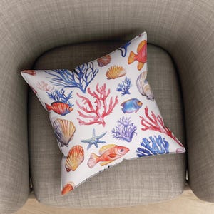 Op de afbeelding: Vierkant kussen met een witte achtergrond en een aquarelpatroon van koraal, schelpen, zeesterren en vissen. Het ontwerp bevat rode, blauwe, oranje en gele elementen. Het kussen ligt op een grijze fauteuil.