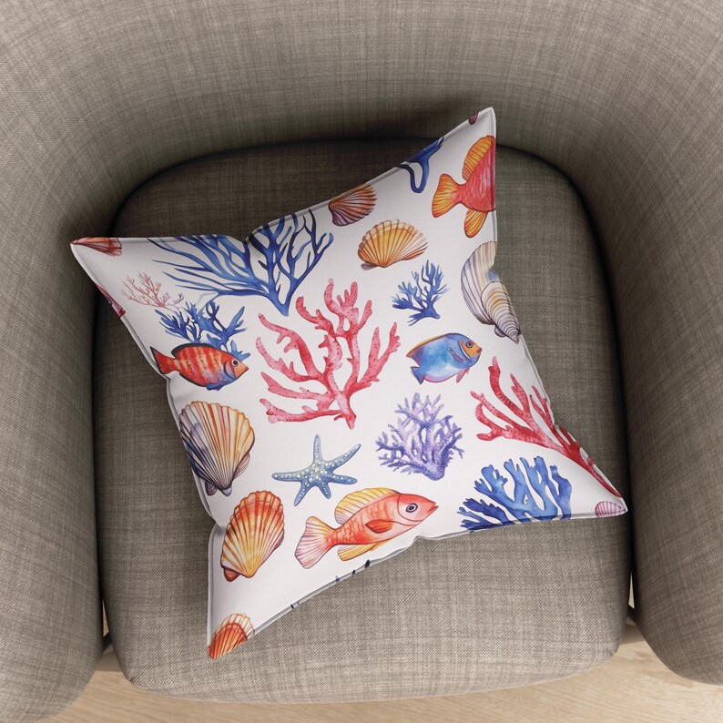 Op de afbeelding: Vierkant kussen met een witte achtergrond en een aquarelpatroon van koraal, schelpen, zeesterren en vissen. Het ontwerp bevat rode, blauwe, oranje en gele elementen. Het kussen ligt op een grijze fauteuil.