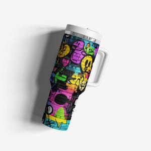 Op de afbeelding: Een reismok met witte handgreep en een kleurrijk graffiti-ontwerp. Het ontwerp toont verschillende cartoongezichten en -vormen in geel, roze, paars en groen op een zwarte bakstenen achtergrond. De mok heeft een wit deksel.