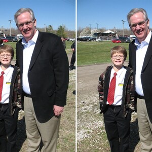 May include: Two men and a boy are standing together. The man on the left is wearing a black suit and a white shirt. The man on the right is wearing a black suit and a white shirt. The boy is wearing a black jacket, a white shirt, a red tie, and blue pants.