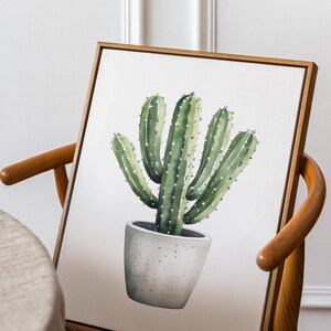May include: A framed watercolor painting of a cactus in a gray pot. The cactus is green with white dots and spines. The frame is a light brown color, and the painting is leaning against a wooden chair.