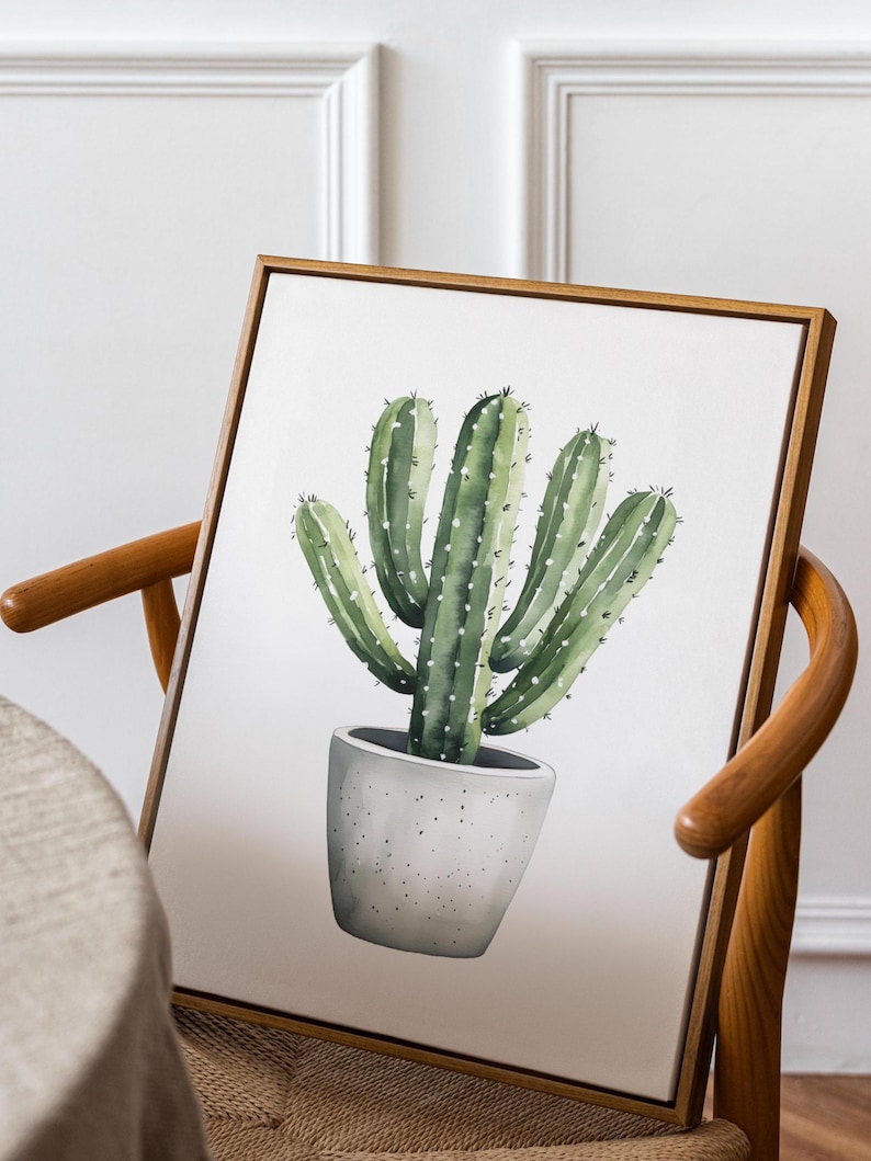 May include: A framed watercolor painting of a cactus in a gray pot. The cactus is green with white dots and spines. The frame is a light brown color, and the painting is leaning against a wooden chair.