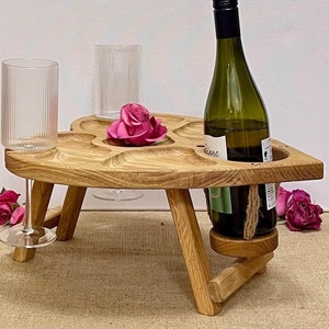 Può includere: Un tavolo da picnic in legno a forma di cuore con una bottiglia di vino, due bicchieri e una rosa rosa. Il tavolo ha incavi per calici e una bottiglia. Il tavolo è in legno chiaro e ha gambe pieghevoli.