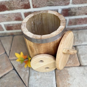 Può includere: Una botte di legno con un coperchio, realizzata in legno naturale con una fascia di metallo argentato attorno alla parte superiore. La botte è appoggiata su una superficie di pietra.