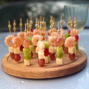 Puede incluir: Una tabla redonda de madera con brochetas de camarones, queso, uvas y kiwi. Las brochetas tienen adornos dorados en la parte superior. Una copa de vino en el fondo.