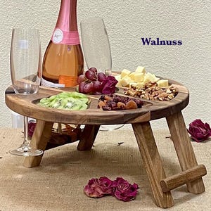 Könnte beinhalten: Ein Picknicktisch aus Holz mit runder Platte und Klappbeinen. Der Tisch enthält eine Flasche Rosé-Champagner, zwei Sektgläser, Trauben, Käse, Nüsse und Kiwischeiben. Das Wort "Walnuss" ist im Hintergrund aufgedruckt.