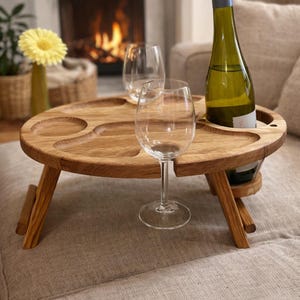 Op de afbeelding: Een ronde inklapbare houten tafel met uitgesneden inkepingen voor snacks en twee ronde uitsparingen voor wijnglazen. Een groene glazen wijnfles staat in een van de openingen en een transparant wijnglas staat op het tafeloppervlak.