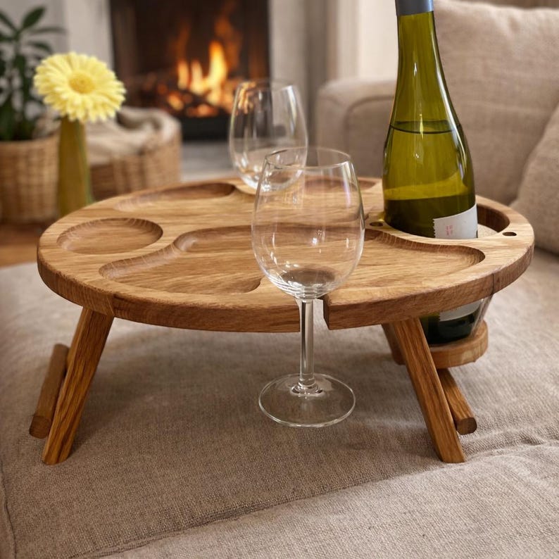 Op de afbeelding: Een ronde inklapbare houten tafel met uitgesneden inkepingen voor snacks en twee ronde uitsparingen voor wijnglazen. Een groene glazen wijnfles staat in een van de openingen en een transparant wijnglas staat op het tafeloppervlak.