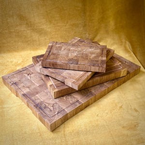 May include: A stack of end-grain butcher block cutting boards. The boards are made of light brown wood and have a square or rectangular shape. Each board has a carved groove around the edge.