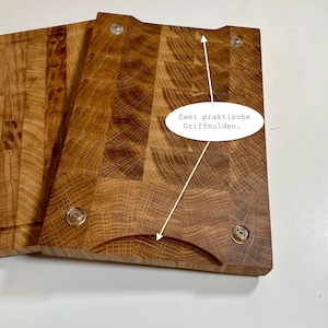 May include: Two end-grain wooden cutting boards with rounded handle cutouts and clear, round feet. The boards are made of light brown wood with a block pattern. The text in the image says "Zwei praktische Griffmulden."