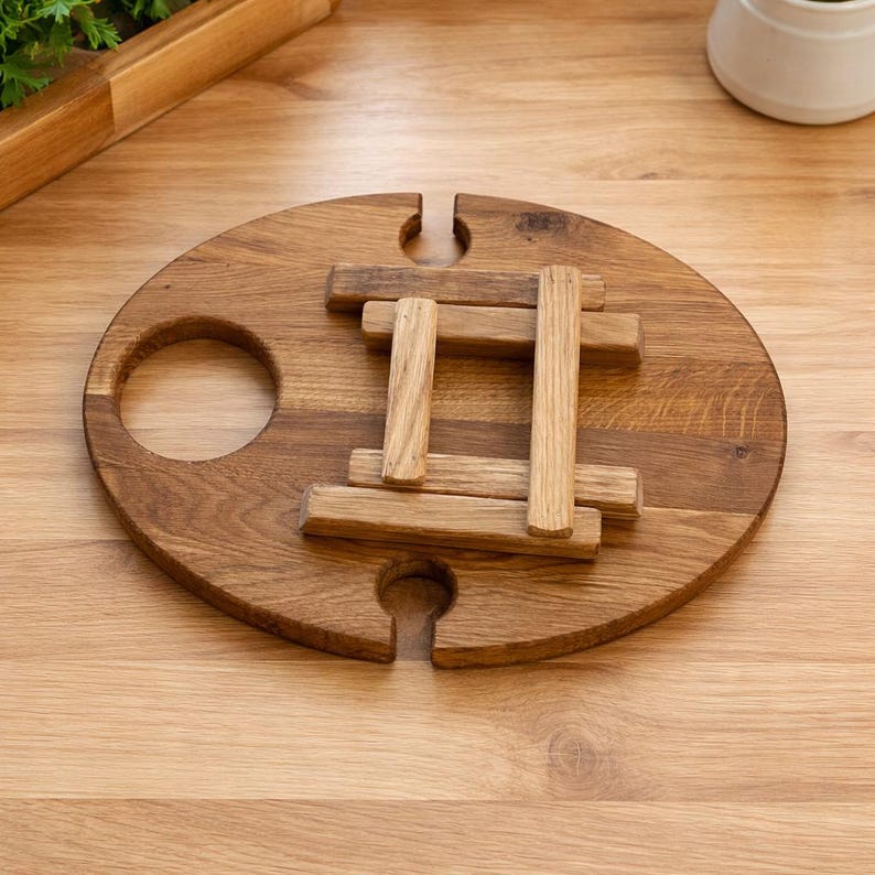 Op de afbeelding: Een ronde picknicktafel van bruin hout met een natuurlijke houtnerfafwerking. De tafel heeft een centrale uitsparing voor een wijnfles en twee kleinere zijdelingse uitsparingen voor glazen, met een inklapbare basis van vier houten latten.