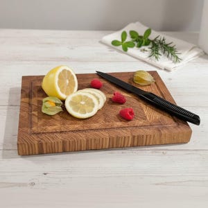 May include: A rectangular wooden cutting board with a juice groove, featuring a black knife, sliced lemons, raspberries, and a green husk tomato. The board is made of light brown wood and is surrounded by a white background.
