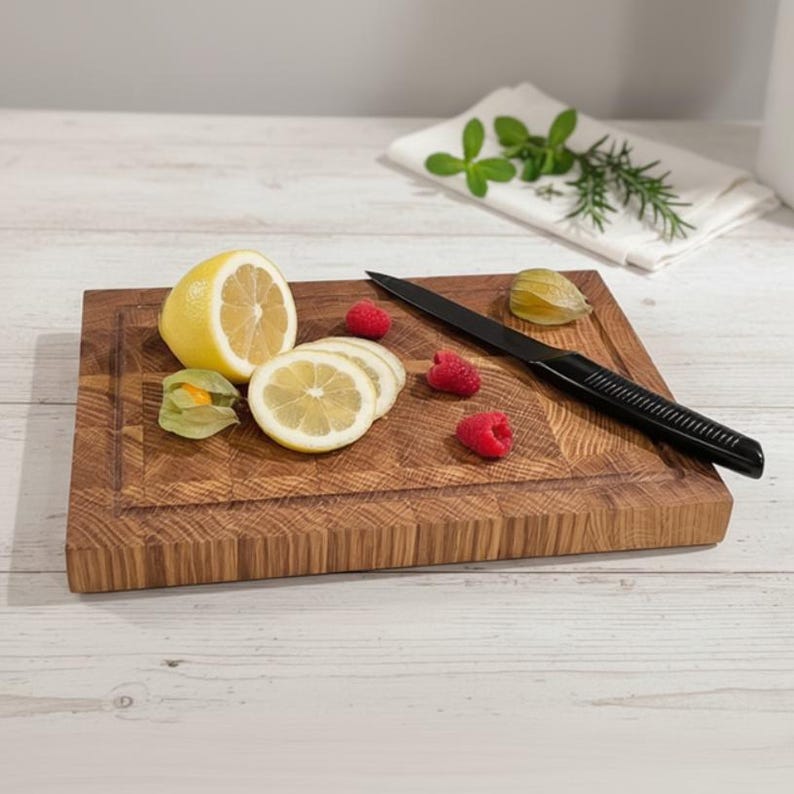 May include: A rectangular wooden cutting board with a juice groove, featuring a black knife, sliced lemons, raspberries, and a green husk tomato. The board is made of light brown wood and is surrounded by a white background.
