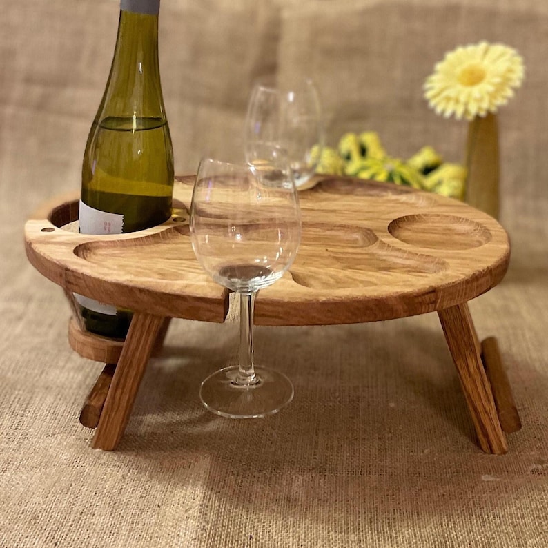 Op de afbeelding: Een ronde houten picknicktafel met inklapbare poten, voorzien van een ronde uitsparing voor een wijnfles en twee extra uitsparingen voor wijnglazen. Een glazen wijnfles en twee lege wijnglazen staan op de tafel geplaatst.