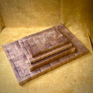 May include: A stack of four wooden cutting boards of varying sizes. The boards are made of light brown wood with a block pattern and a dark border. The boards are arranged from largest to smallest, creating a layered effect.