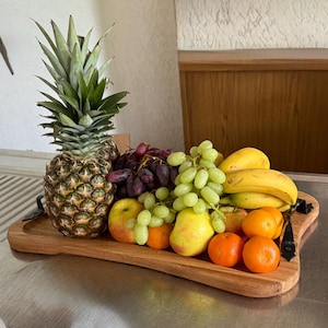 Peut inclure: Un plateau de service en bois rempli de fruits frais. On y trouve un ananas, des grappes de raisins verts et violets, des bananes, des pommes, des poires et des mandarines. Le plateau est orné de détails métalliques.