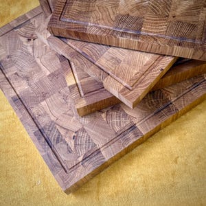 May include: A stack of end-grain butcher block cutting boards. The boards are made of light brown wood with a square pattern and a recessed border. The boards are of varying sizes and are stacked on top of each other.