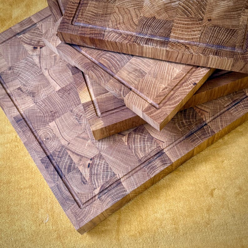 May include: A stack of end-grain butcher block cutting boards. The boards are made of light brown wood with a square pattern and a recessed border. The boards are of varying sizes and are stacked on top of each other.