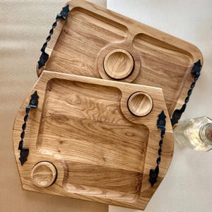 Könnte beinhalten: Zwei Holzserviertabletts mit natürlicher Holzmaserung. Jedes Tablett hat eine Aussparung, eine kreisförmige Vertiefung und eine kleinere kreisförmige Vertiefung. Schwarze Metallgriffe und Akzente zieren die Seiten. Im Hintergrund ist eine Glasflasche zu sehen.