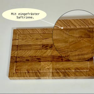 May include: A rectangular wooden cutting board with a juice groove. The board is made of light brown wood and has a textured surface. The text "Mit eingefr&auml;ster Saftrinne" is in a yellow oval.