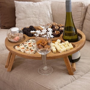 Op de afbeelding: Een ronde houten snacktafel met inklapbare poten en uitgesneden vakken gevuld met kaasblokjes, gedroogde abrikozen, stervormige koekjes en pure chocoladelekkernijen, inclusief een fles witte wijn en twee lege wijnglazen op de tafel.