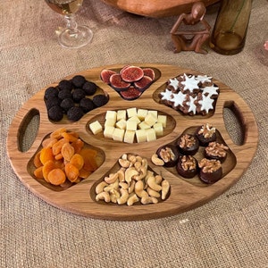 Könnte beinhalten: Ein Holztablett mit sieben separaten Fächern, die jeweils mit verschiedenen Snacks gefüllt sind. Die Fächer enthalten getrocknete Aprikosen, Cashews, Schokoladen-Walnüsse, Käsewürfel, Feigen, sternförmige Kekse und Schokoladen-Trüffel.