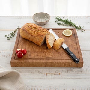 May include: A wooden cutting board with a loaf of sliced bread, a bread knife, and a small bowl. The bread is golden brown, and the knife has a black handle. Red peppers and a lemon slice are also on the board.