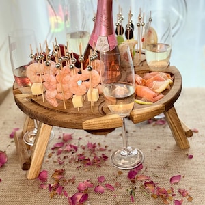 Può includere: Un tavolo da picnic in legno con un piano rotondo e due gambe su ogni lato. Il tavolo ha un porta bottiglie integrato e quattro porta bicchieri. Il tavolo è apparecchiato con una bottiglia di vino rosato, due bicchieri di vino, spiedini di gamberi, cubetti di formaggio e salmone affumicato.