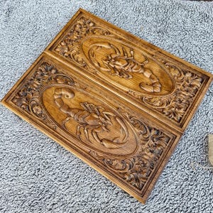 Könnte beinhalten: Backgammon-Spielbrett aus hellem Holz mit einem geschnitzten Skorpion-Design. Das Brett ist in zwei Hälften geteilt, jede mit einem ovalen Skorpion-Schnitzwerk, umgeben von aufwendigen Schnörkeln. Das offene Brett zeigt die Spielfläche.