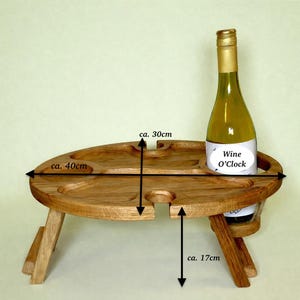 Op de afbeelding: Een bruine inklapbare houten picknicktafel met een ovaal blad voorzien van uitsparingen voor glazen en een fles. De tafel is ongeveer 40 cm breed, 30 cm diep en 17 cm hoog. Een wijnfles met het etiket Wine O'Clock staat in een van de ronde openingen.