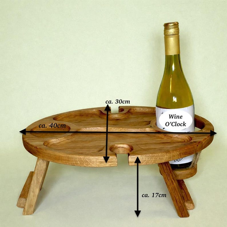 Op de afbeelding: Een bruine inklapbare houten picknicktafel met een ovaal blad voorzien van uitsparingen voor glazen en een fles. De tafel is ongeveer 40 cm breed, 30 cm diep en 17 cm hoog. Een wijnfles met het etiket Wine O'Clock staat in een van de ronde openingen.