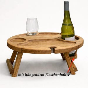 Op de afbeelding: Een ronde inklapbare houten tafel met uitgesneden inkepingen voor een glas en een fles. De tafel heeft een hangende flessenhouder aan de zijkant, aangegeven door een rode pijl en de Duitse tekst "mit h&auml;ngendem Flaschenhalter".